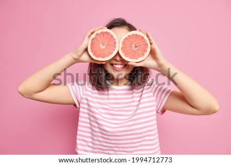 Happy brunette woman covers eyes with fresh juicy grapefruit slices smiles gladfully keeps to healthy diet wears casual striped t shirt isolated over pink background. Tropical fruit contaning vitamins Royalty-Free Stock Photo #1994712773