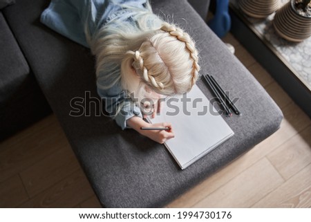 Preschooler girl laying at the sofa and pondering while drawing at the paper