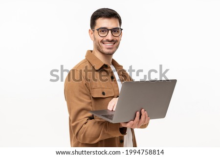 Portrait of young modern business man standing in casual brown shirt, holding laptop and looking at camera with happy smile, isolated on gray