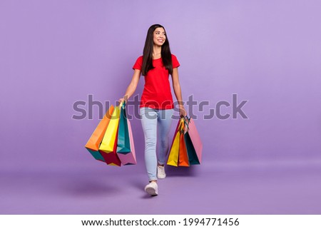 Photo of shiny sweet woman dressed red t-shirt looking empty space walking holding colorful bargains isolated purple color background