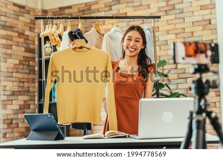 Young and beautiful Asian woman blogger showing clothes in front of smartphone camera while recording vlog video and live streaming at her shop Royalty-Free Stock Photo #1994778569