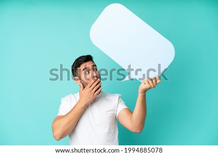 Young handsome caucasian man isolated on blue background holding an empty speech bubble with surprised expression