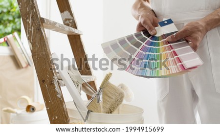hands of house painter man decorator choose the color using the sample swatch, work of the house to renovate, a wooden ladder with paint brushes and a bucket as a background, close up Royalty-Free Stock Photo #1994915699