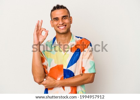 Young venezuelan man isolated on white background winks an eye and holds an okay gesture with hand.