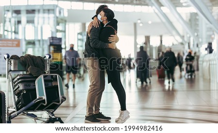 Woman in face mask receiving man at airport arrival. Couple meeting and hugging each other at airport arrival gate. Royalty-Free Stock Photo #1994982416