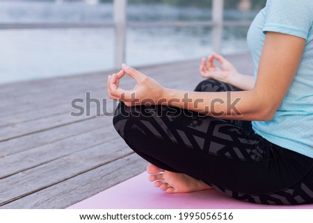 Cropped shot of a young woman who meditates while holding fingers in yoga sign sitting on the sea pier. Peaceful girl keeps hands in mudra gesture sitting in the lotus pose