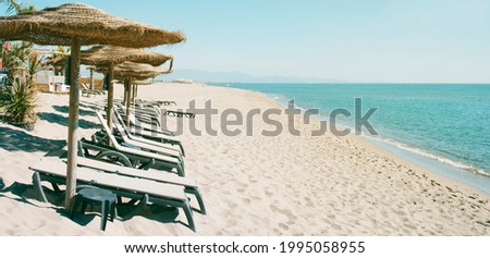 some sun umbrellas made with natural materials and some beige sunloungers, on a quiet beach, in a panoramic format to use as web banner or header