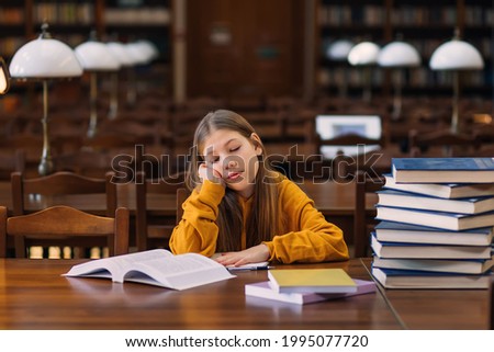 cute exemplary schoolgirl smiling while sitting with books at the table in the library or classroom, ready for a lesson.