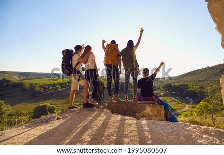 Back view of company of hikers with backpacks celebrating successful achievement while standing with raised arms on hill at sunset during summer adventure Royalty-Free Stock Photo #1995080507