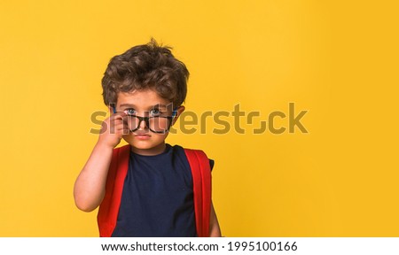 Cute little nerd smart boy wearing glasses isolated on yellow background. Funny pupil with red big backpack ready to go to school. Confident child looking to camera.