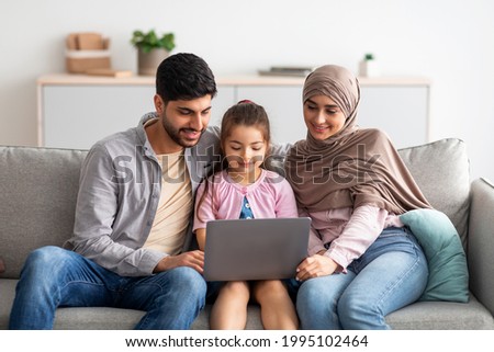 Cheerful muslim family having fun with laptop, surfing internet, playing games or having video chat, sitting together on sofa. Happy arab man, child and woman in hijab resting at home