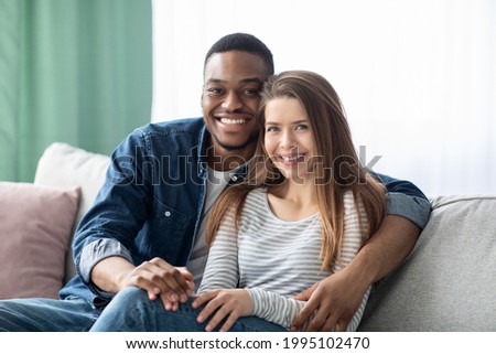 Portrait Of Smiling Young Multicultural Couple Posing In Home Interior, Romantic Interracial Lovers Embracing And Looking At Camera While Relaxing On Couch In Cozy Living Room, Enjoying Time Together