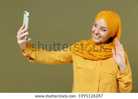 Young friendly arabian asian muslim woman 20s in abaya hijab yellow clothes do selfie shot on mobile phone waving hand greeting isolated on olive green background People uae islam religious concept
