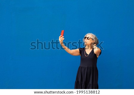 Senior grey haired woman taking a selfie with a red smartphone against a blue wall