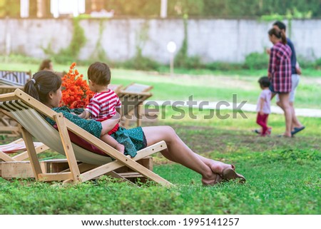Back view of Asian mom and son sitting on garden chair and by the table in garden. Summer vacation in green surroundings. Happy person outdoors relaxing on deck chair in garden. Outdoor leisure.