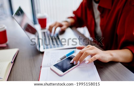 Crop faceless woman remote worker in casual clothes sitting at table surfing mobile phone and typing on laptop while working on project