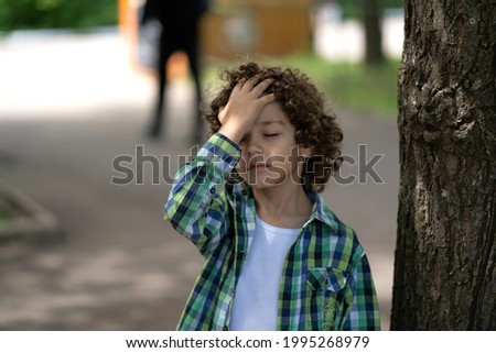 A boy with curly hair stands in the street and holds on to his forehead. Baby got sunstroke Royalty-Free Stock Photo #1995268979
