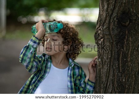 A little boy with curly hair stands in the street and puts a bottle of water to his forehead. Baby got heatstroke Royalty-Free Stock Photo #1995270902