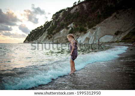 A girl in a purple sundress stands on the surf line against the background of the sea and rocks