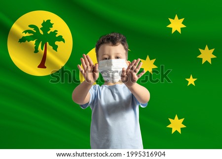 Little white boy in a protective mask on the background of the flag of Cocos Islands. Makes a stop sign with his hands, stay at home Cocos Islands.