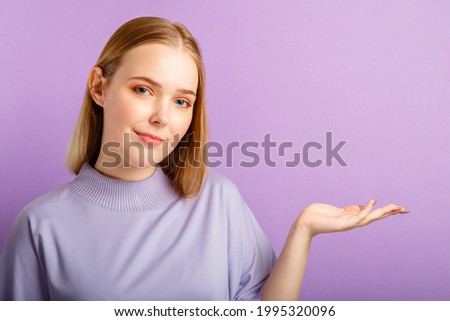 Confident blonde caucasian pretty young woman show by hand on copy space place over isolated color purple background.