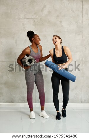 Happy African American sportswoman and her Caucasian female friend having fun and laughing against the wall at health club. Copy space.  Royalty-Free Stock Photo #1995341480
