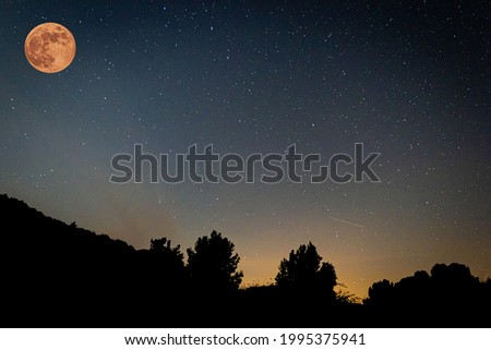 The strawberry supermoon in the dark night in the sky gradient from orange to blue over silhouettes of mountains and trees in June 2020 in Spain. Royalty-Free Stock Photo #1995375941