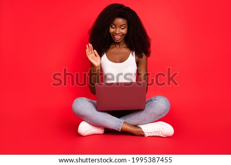 Full size photo of young afro girl happy positive smile sit floor speak video call laptop wave hi isolated over red color background