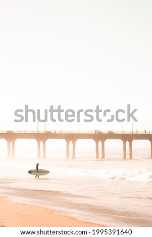 Bright and light early morning surf picture, with a surfer, waves, and a pier. Light oranges and yellows give a beautiful, joyful, and lighthearted feel to this picture.