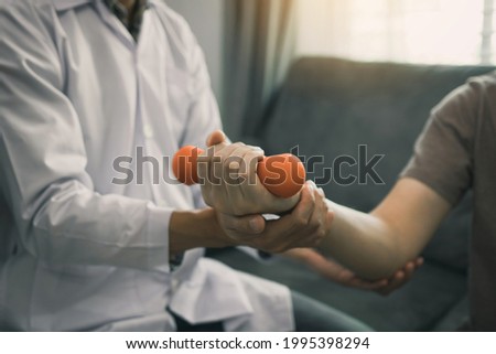 Asian young male physiotherapist helping patient with lifting dumbbells exercises in office. Royalty-Free Stock Photo #1995398294