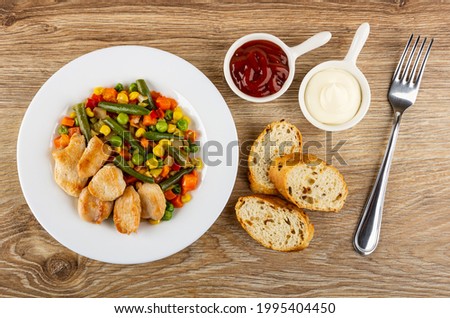 White glass plate with pieces of fried chicken meat and vegetable blend, sauce boats with tomato ketchup and mayonnaise, slices of bread, fork on wooden table. Top view Royalty-Free Stock Photo #1995404450