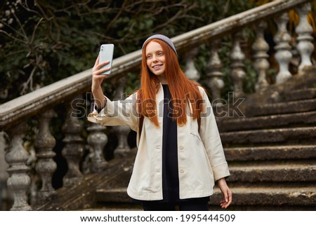 Portrait Of Cute Modern Woman Taking Selfie, Photo On The Background Of Building Stairs, Historical Ancient Place, Side View. Optimistic Cheerful Lady In Casual Coat And Hat, With Backpack