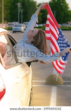 Young millennial blonde woman looking from car with american flag. Flag of the united states in her hands. July 4th Independence Day. USA patriotism national holiday. Caucasian girl. Copy space. 