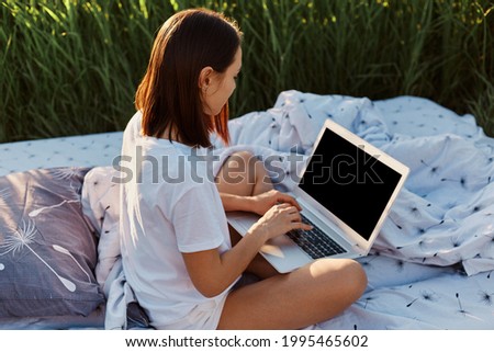 Side view of dark haired woman wearing white casual T-shirt sitting with crossed legs and holding lap top with blank screen, freelancer working online on soft bed in meadow.