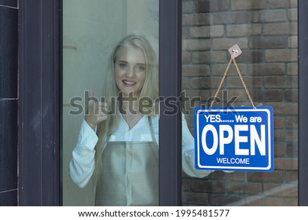 Beautiful caucasian woman with blonde hair, shop's owner standing with confidence in front of shop with open sign, business reopen after Coronavirus quarantine is over.