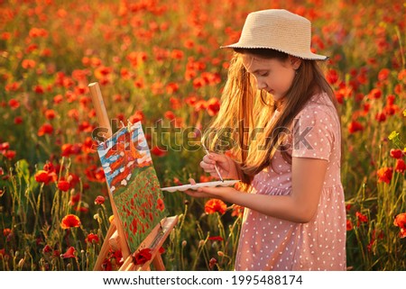 Little girl is standing in the field of red poppies and painting on the canvas placed on a drawing stand. Be a part of learning outside of the school in the nature park. Creative hobby