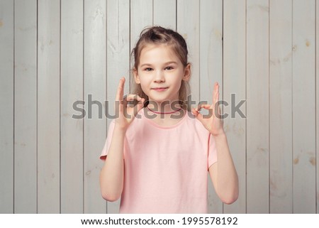 Okay deal. Smiling good-looking little girl 9-11 years old showing her support, make OK sign, say yes, agree and approve, guarantee quality, standing in casual pink t shirt over wooden background
