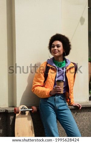 Happy african american woman with coffee to go standing near longboard and facade of building