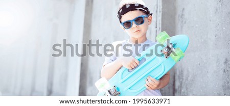 Stylish boy child in street clothes in a T-shirt, bandana, blue sunglasses and black and white sneakers with a turquoise skateboard in his hands against the background of a concrete wall.