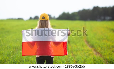 Blond girl holding flag of Poland in a green field. Back view. Polish Flag Day. Independence Day. Travel and learn polish language concept. Royalty-Free Stock Photo #1995635255