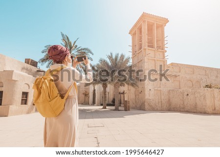 Happy woman wearing a turban hat takes pictures on her smartphone against the background of a Bur Dubai old town near Creek district