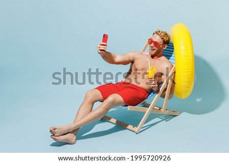 Full length young man in red shorts swimsuit glasses sit in chair inflatable ring do selfie shot mobile cell phone hold cocktail isolated on pastel blue background Summer vacation sea sun tan concept