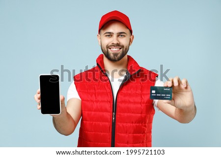 Delivery guy employee man in red cap white T-shirt uniform workwear work as dealer courier hold use mobile cell phone credit bank card isolated on pastel blue color background studio. Service concept