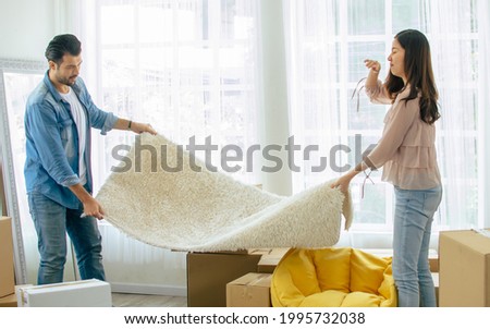 Lovely sweet Caucasian and Asian couple helping each other to decorate their home by taking carpet together. Woman has allergy of dust and moving to new comfortable house or apartment with happiness