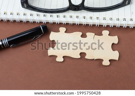Part of jigsaw puzzle on the brown background with a pen, and eyeglasses on the notebook