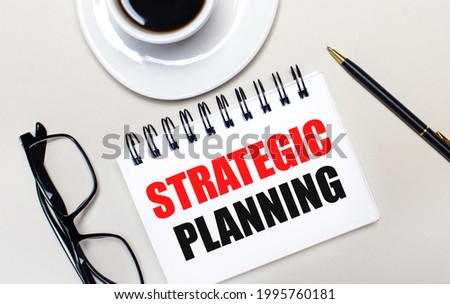 Glasses, a white cup of coffee, a white notebook with the words STRATEGIC PLANNING and a ballpoint pen lie on a light background. Flat lay. View from above.
