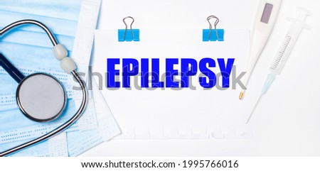 On a light background lie a stethoscope, an electronic thermometer, a syringe, a face mask and a sheet of paper with the text EPILEPSY. Medical concept