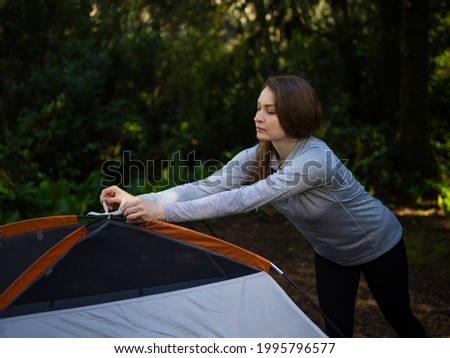 In the photo we see a young woman at the weekend. The woman lays out the tent. In the photo, we also see a dense green forest in the background. Relax, healthy lifestyle. Blank space to insert