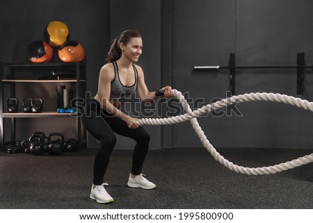 Muscular young smiling woman working out with battle ropes at dark gym, intense functional circuit training. Gray gym background with sports equipment. Crossfit, fitness and workout concept Royalty-Free Stock Photo #1995800900