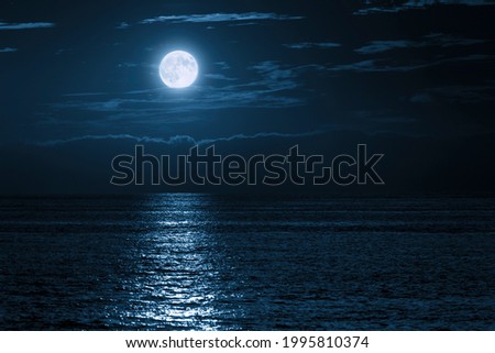 This large full blue moon rises brightly over the cloud bank in this calm ocean creating sparkles accross the waves in this beautiful tranquil scene.  Royalty-Free Stock Photo #1995810374